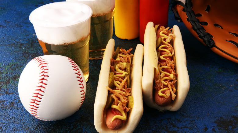 Two stadium hot dogs sit near plastic cups of beer, a baseball, a baseball glove, and ketchup and mustard dispensers