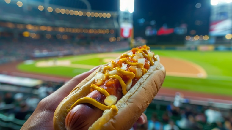 Hand holding hot dog with baseball stadium in background