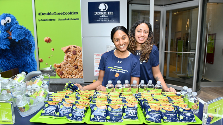 Two people serving allergen-free chocolate chip cookies and water bottles outside of a Double Tree by Hilton hotel with a sign that has Cookie Monster on it