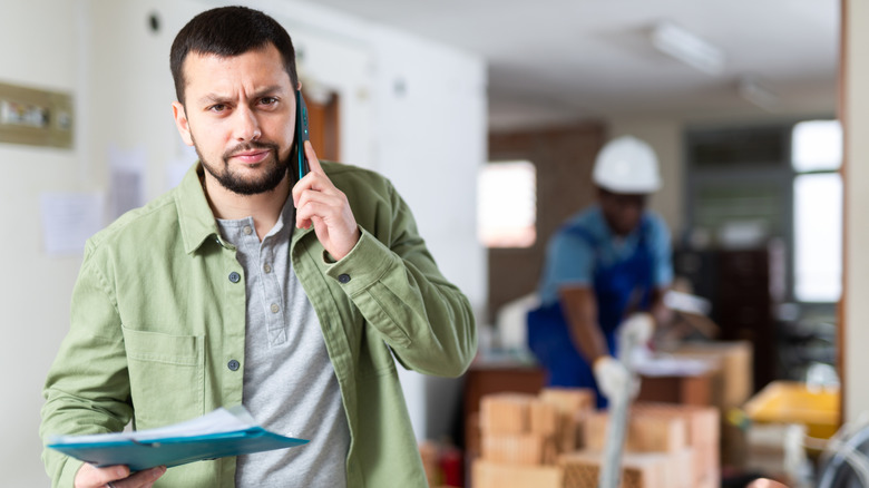 A DIYer with a frustrated expression holds documents while talking on a cellphone, as a person with a hard hat works in the background