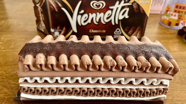 Viennetta accordion-like layered chocolate and vanilla ice cream cake in front of box, on a wood table. Blurred trinkets in the background.
