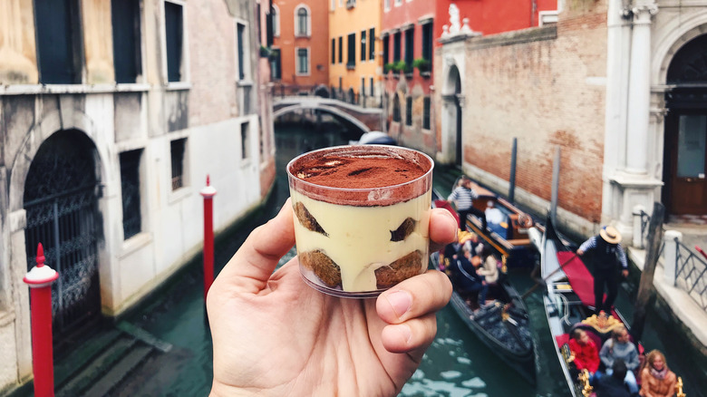 A hand holding up a tiramisu above a Venetian canal
