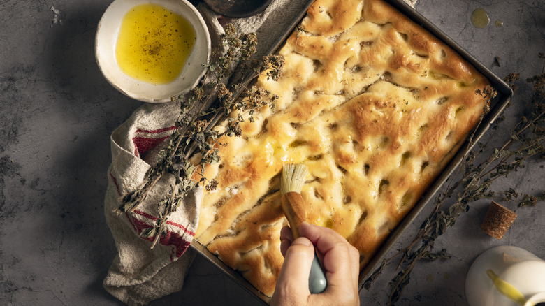 Fresh focaccia next to a bowl of olive oil and rosemary sprig with a hand brushing it with olive oil.