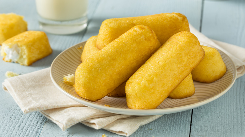 Several Twinkies sit on a plate, with a glass of milk and a Twinkie broken in half in the background