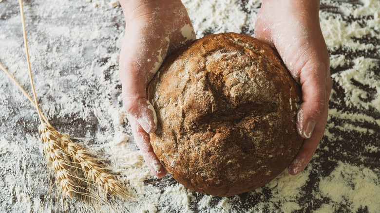 Hands holding wheat bread loaf over floured surface