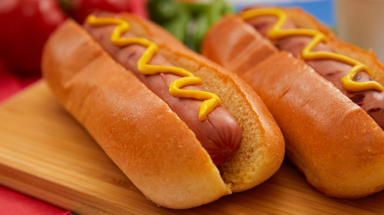 A small hot dog covered in mustard sits in the middle of a large, fluffy bun
