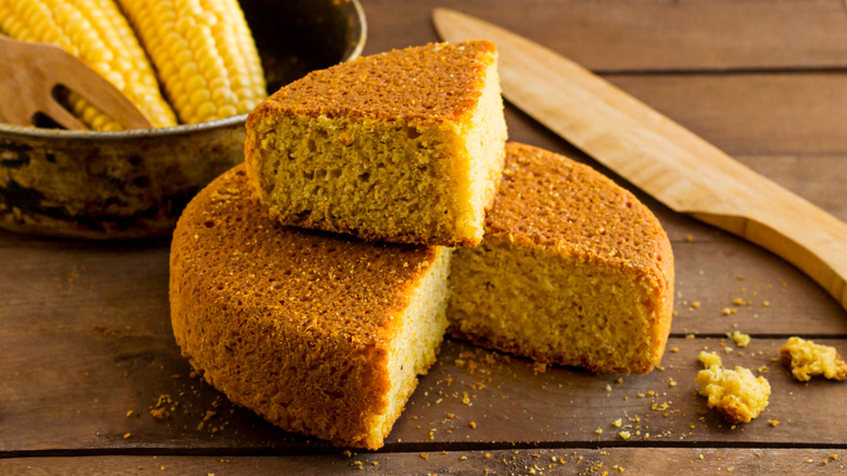 Cornbread with one slice removed and placed atop the loaf next to a wooden knife and pot of corn