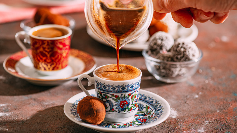 Hand pouring Turkish coffee into a small, decorated coffee cup with a chocolate truffle on the saucer. A second filled cup and other truffles are blurred behind, and all on a texturized, sandy table.