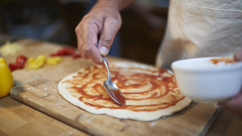 close up of a hand spreading sauce on pizza dough with a spoon