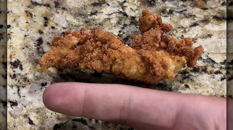 A person holds up one finger against a Chick-fil-A chicken tender for a size comparison