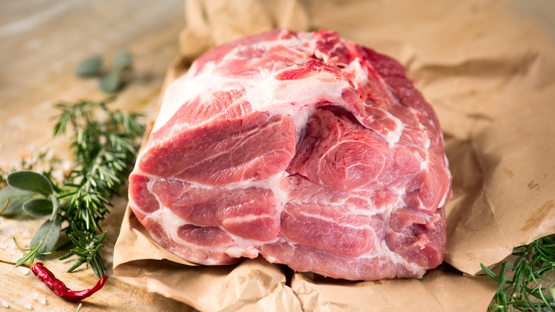 A raw chunk of pork sits on butcher paper with chili peppers and herbs