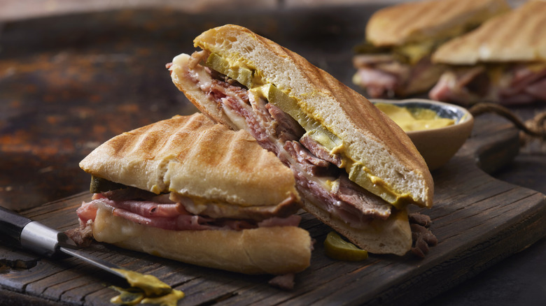A Cuban sandwich cut in half with a mustard-smeared knife on the cutting board