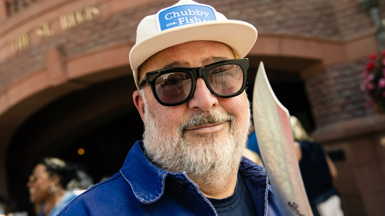 Andrew Zimmern wears sunglasses and a hat and smiles.