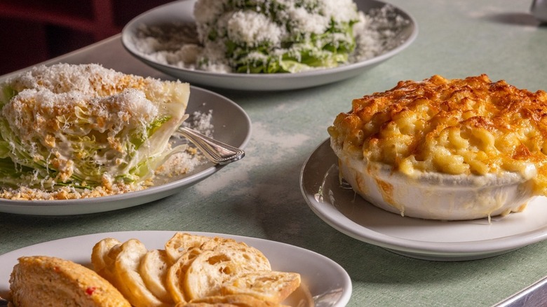 A golden-topped dish of mac and cheese sits on a bar with two salads and a bread bowl.