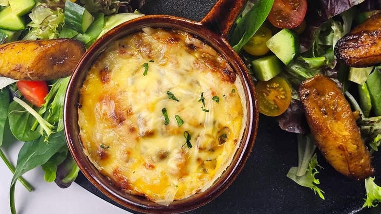 Dish of traditional Keshi Yena cheese dish surrounded by veggies and cooked plantains