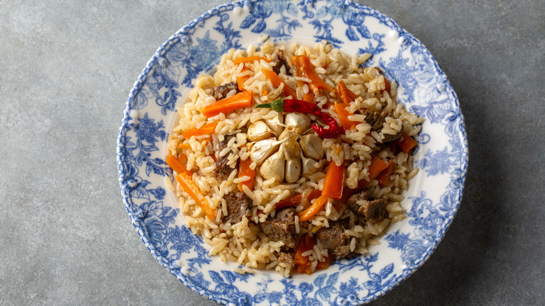 Blue and white bowl of Uzbek plov with rice, veggies, meat, and roasted garlic cloves