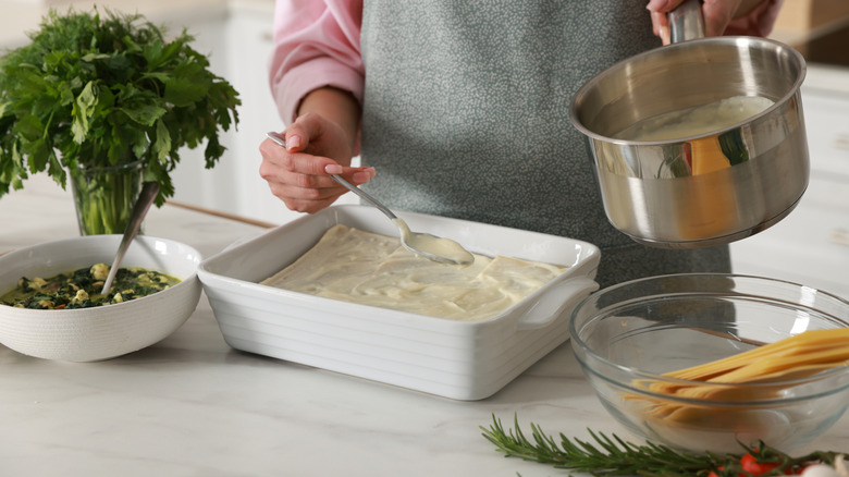 A cook spoons béchamel sauce onto a white lasagna next to herbs and uncooked pasta