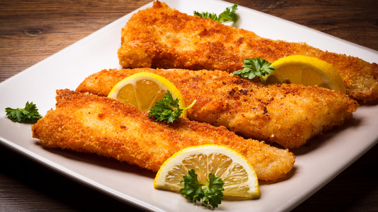 Three pieces of golden fried fish on a white plate garnished with lemon slices and parsley.