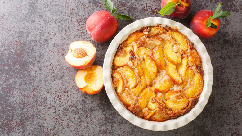A peach cake rests in a cake pan next to whole and sliced peaches