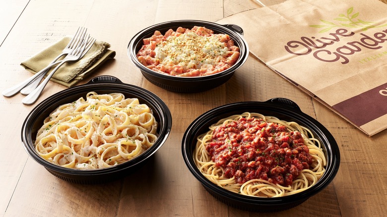 Three disposable bowls of pasta, featuring spaghetti, fettuccini alfredo, and ziti sit on a table next to a to-go bag
