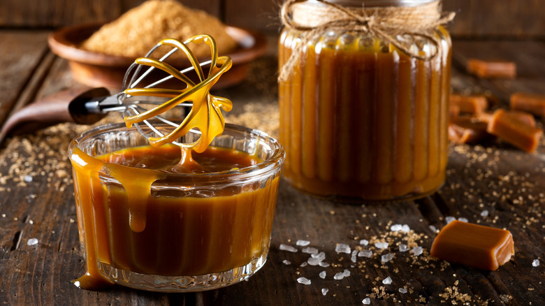 Two ramekins of caramel sauce on a wooden table with a whisk dripping with the sauce and chunks of rock salt scattered on the wood