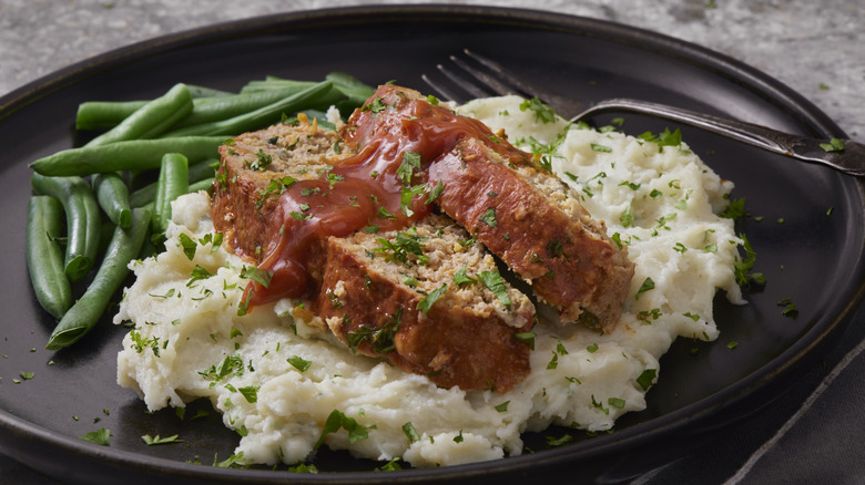 Two slices of turkey meatloaf sit on a bed of creamy mash, topped with fresh herbs, ketchup, and with a side of greenbeans
