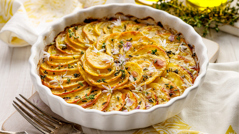 Scalloped potatoes in a white baking dish sit on a white and yellow cloth with a fork lying adjacent