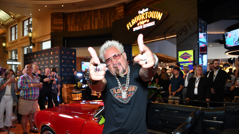 Guy Fieri in front of his red car and Flavortown restaurant pointing his fingers at the camera