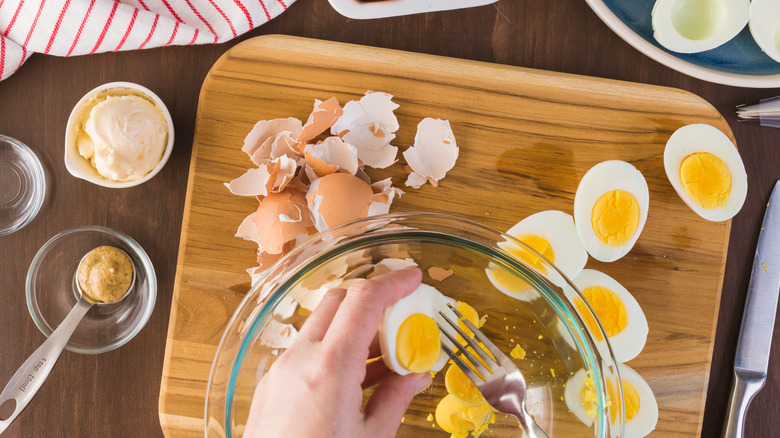 hands preparing deviled eggs with egg shells and other ingredients in background