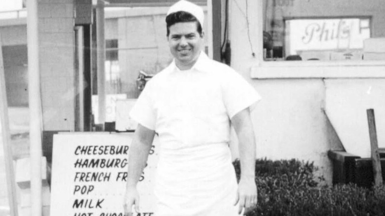 Black-and-white photo of Bob Lang Sr. wearing his In-N-Out uniform standing next to a menu sign