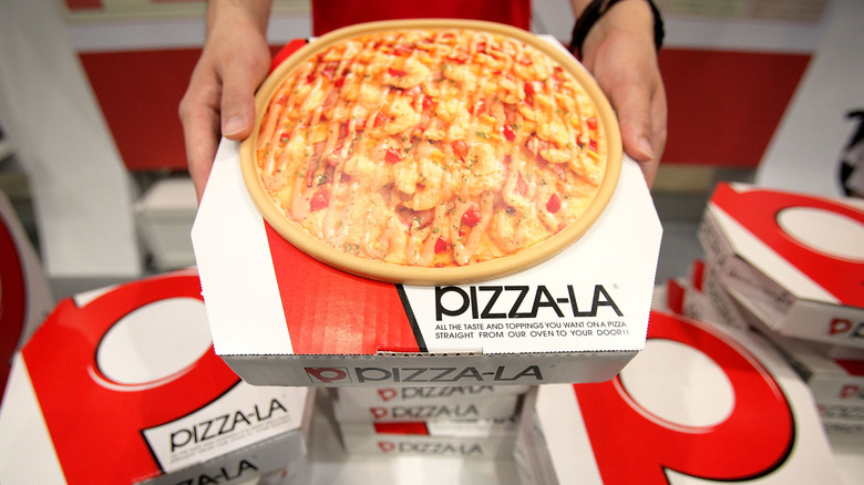 Pizza on top of a Pizza-la box in Japan with hands holding it, and surrounded by additional Pizza-la boxes.