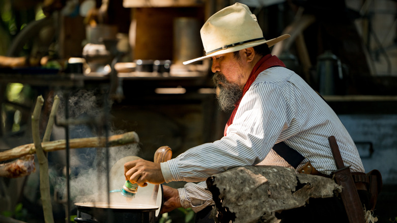 A modern-day cowboy, or one in era-appropriate gear, harkening back to the 19th century, cooks food over an open flame