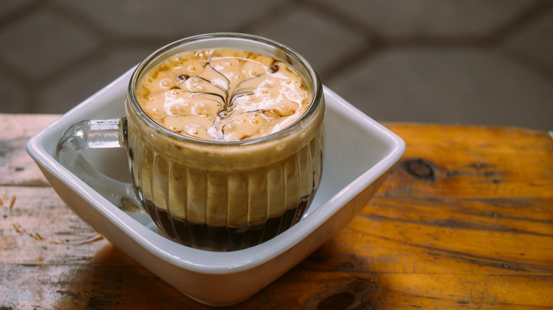 Vietnamese egg coffee in a clear, glass mug in a white bowl on a wooden table.