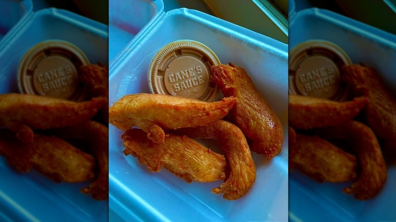 Four naked Raising Cane's chicken tenders in a container with a cup of Cane's Sauce