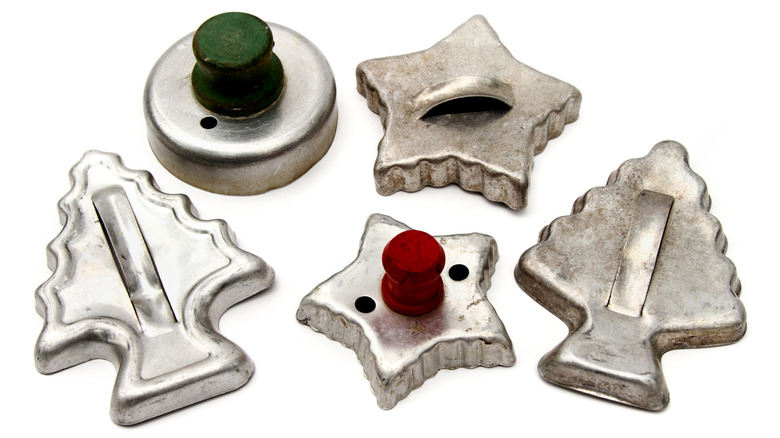 Several vintage cookie cutters, including one with a red wooden top and one with a green wooden top, sit against a white background