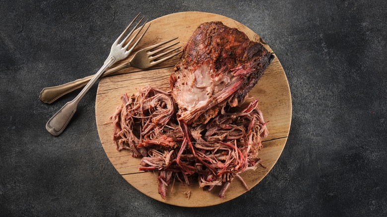 A pile of smoky pulled pork next to a pork shoulder on a circular wooden tray next to two forks