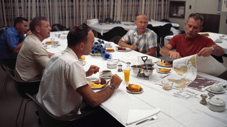 Three Apollo 11 astronauts and two other men eating steak and eggs at long dining table with white tablecloth