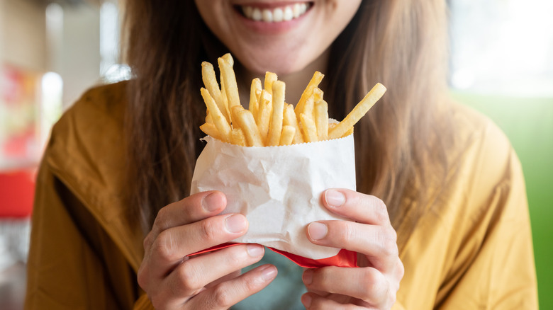A smiling woman holds up a white paper bag of fast food french fries