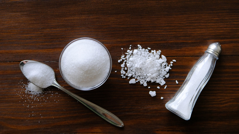 A silver spoon filled with salt, a glass dish of salt, a pile of rock salt, and a salt shaker tipped on its side sit on a wooden surface