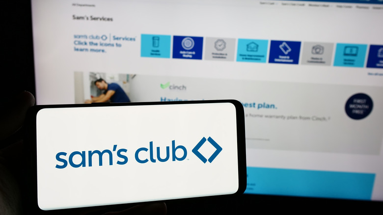 A hand holds up a phone with the Sam's Club app on it, next to a laptop open to the Sam's Club website