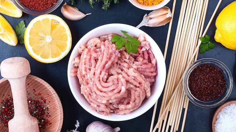 Bowl of ground turkey next to small bowls of spices, wooden skewers, and lemons