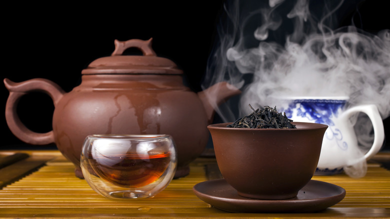 Tea ceremony setting with bowl of loose lapsang souchong black tea, glass of tea, clay teapots and steam