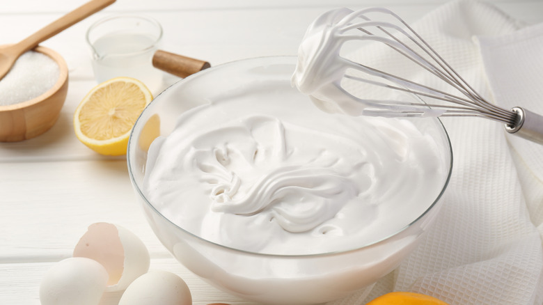 Clear bowl filled with whipped cream and a hand whisk over top, with eggs, lemon, sugar and other ingredients all on a white table.