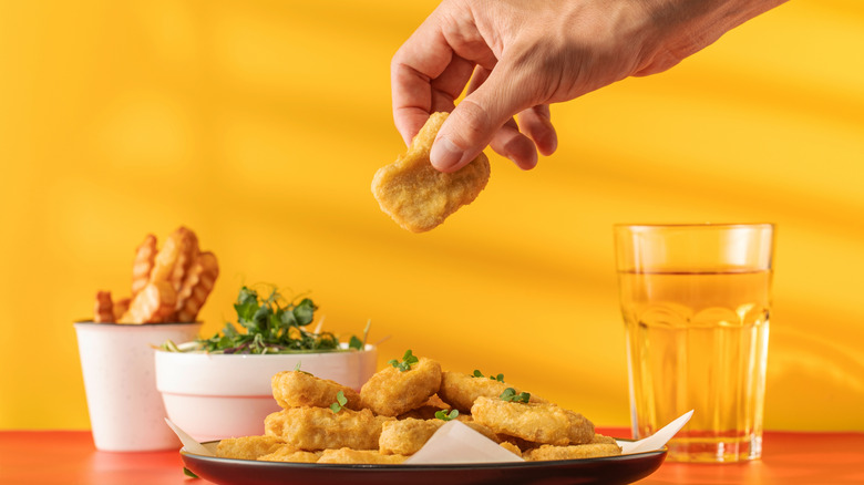 A hand lifts a chicken nuggets off a plate of against a yellow background with bowls of fries and salad and a cup of water