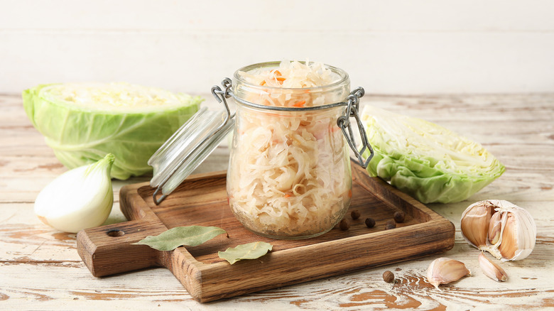 Homemade saerkraut in a jar on a wooden cutting board alongside cabbage, onion, bay leaves, and garlic.