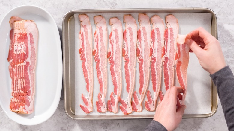 hands putting raw bacon on parchment lined sheet pan