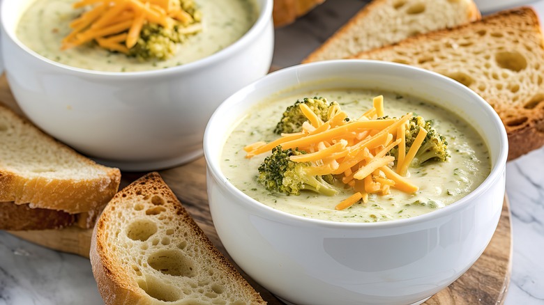 Two bowls of broccoli cheddar soup on a chopping board with sliced bread.