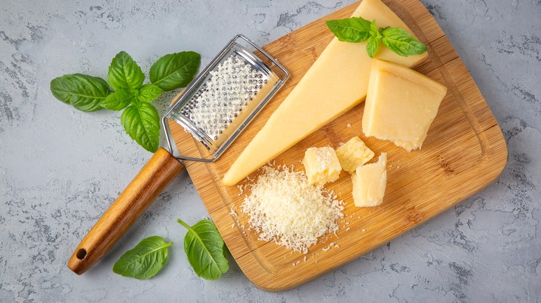 A wedge of cheese on a cutting board with basil, a grater and grated cheese.