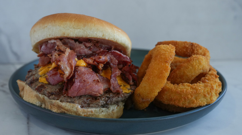 Pastrami spills off a burger served on a bun with onion rings on a blue plate