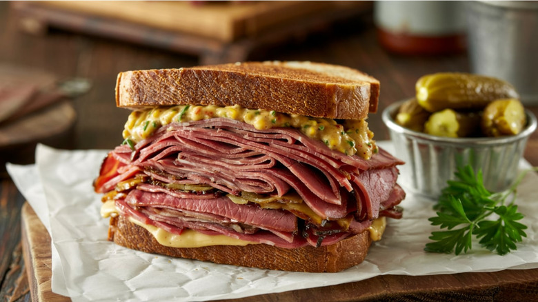 Pastrami stacked high between two slices of bread with mustard served on paper beside pickles